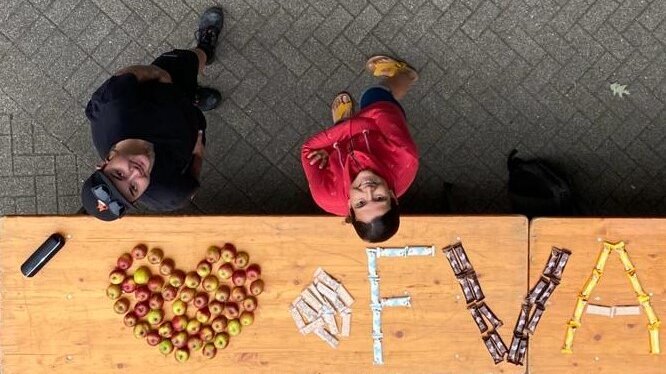 DasBild von Oben Gesundheitsmanagement-Team: Christiane und Alexandra stehe vor einem Tisch, wo sie mit Obst FVA geschrieben haben. Die Kolleginnen schauen nach oben zur Kamera