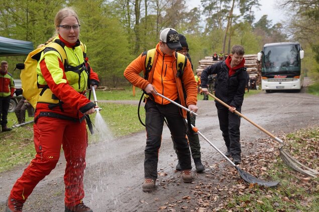 Mehrere Personen nutzen eine Feuerpatsche und schlagen sie auf den Waldboden. Eine Frau im Vordergrund hat außerdem einen Wasserschlauch in der Hand, aus dem Wasser spritzt. 